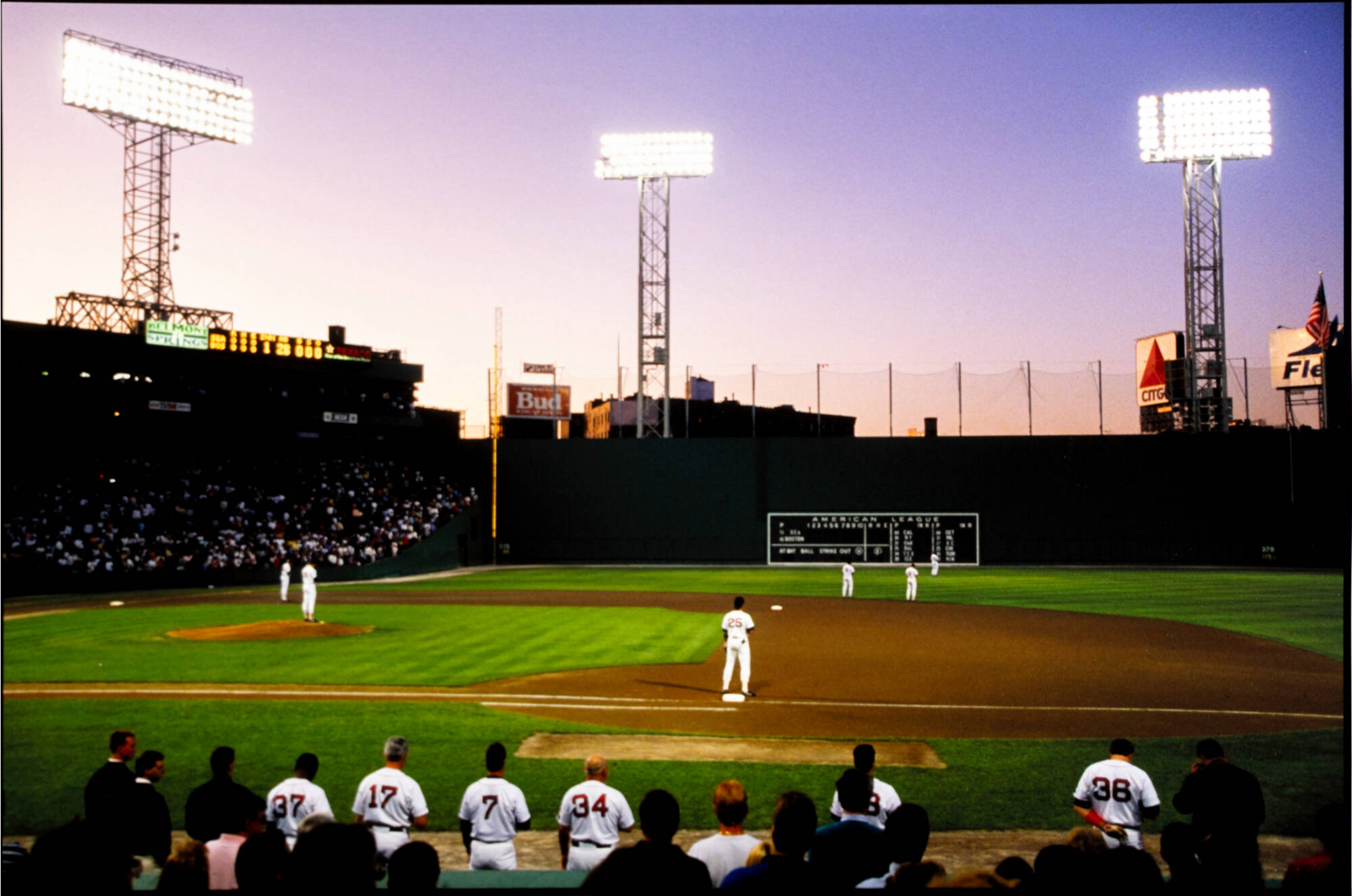 A horizontal color photograph of a baseball stadium. The stadium is full of people and there are players on the field. The sun is setting on the left side of the photo.